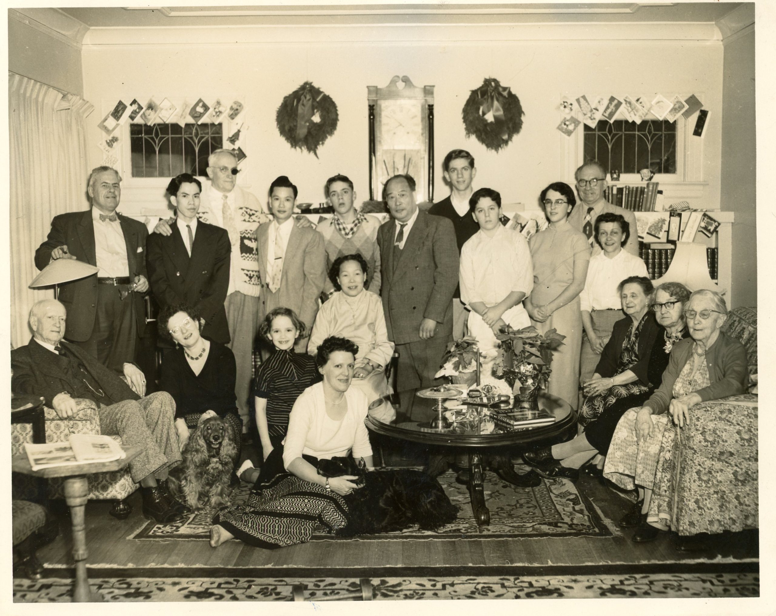 A large group of people, including men, women, and children, pose together in a living room decorated with holiday cards and wreaths. Some are seated while others stand behind them, and a dog sits in front of the group.