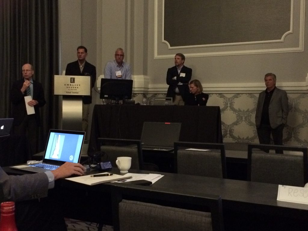 Six people stand and sit at the front of a conference room, some behind a podium and table with computers. Attendees sit facing them, with laptops and notebooks on the tables. The room has gray walls and dim lighting.
