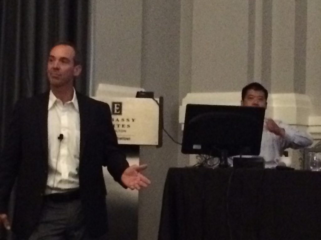 A man in a suit speaks at the front of a room, gesturing with one hand, while another man sits behind a computer at a table. A sign reading Embassy Suites is visible in the background.