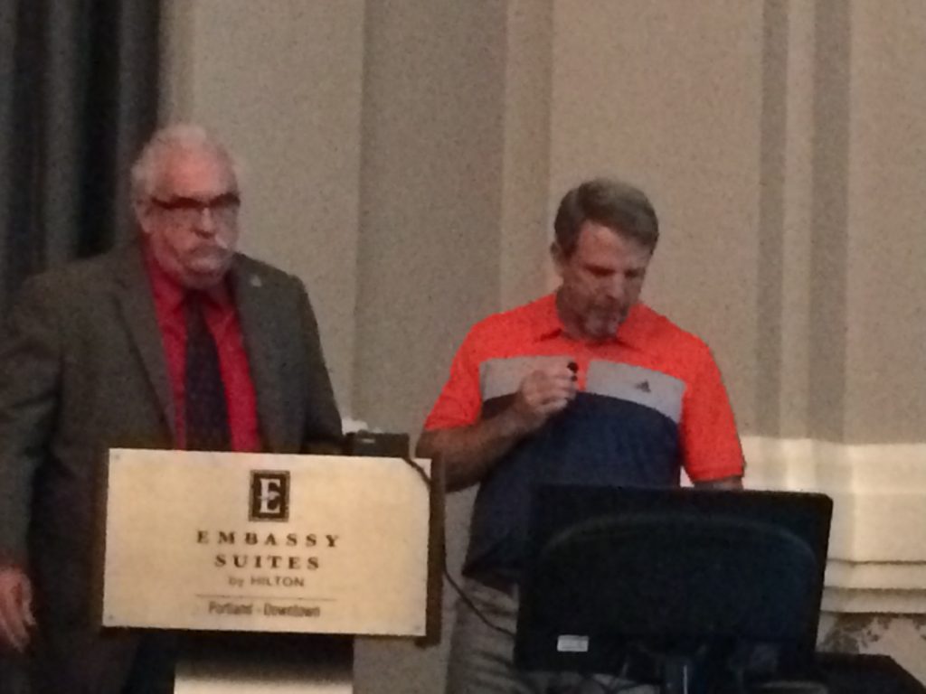 Two men stand at a podium labeled Embassy Suites by Hilton, Portland - Downtown. One wears a brown suit and red shirt, the other an orange and blue polo, looking at a computer screen while holding a microphone.