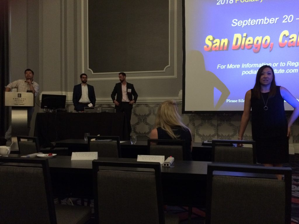People stand at the front of a conference room with a projector screen displaying San Diego, CA. Some sit at tables facing the presenters. The event appears to be a professional seminar or conference.