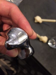 A hand holds a shiny, metallic, anatomically shaped object resembling a knee implant. In the background, two bone-like models are visible on a dark surface.
