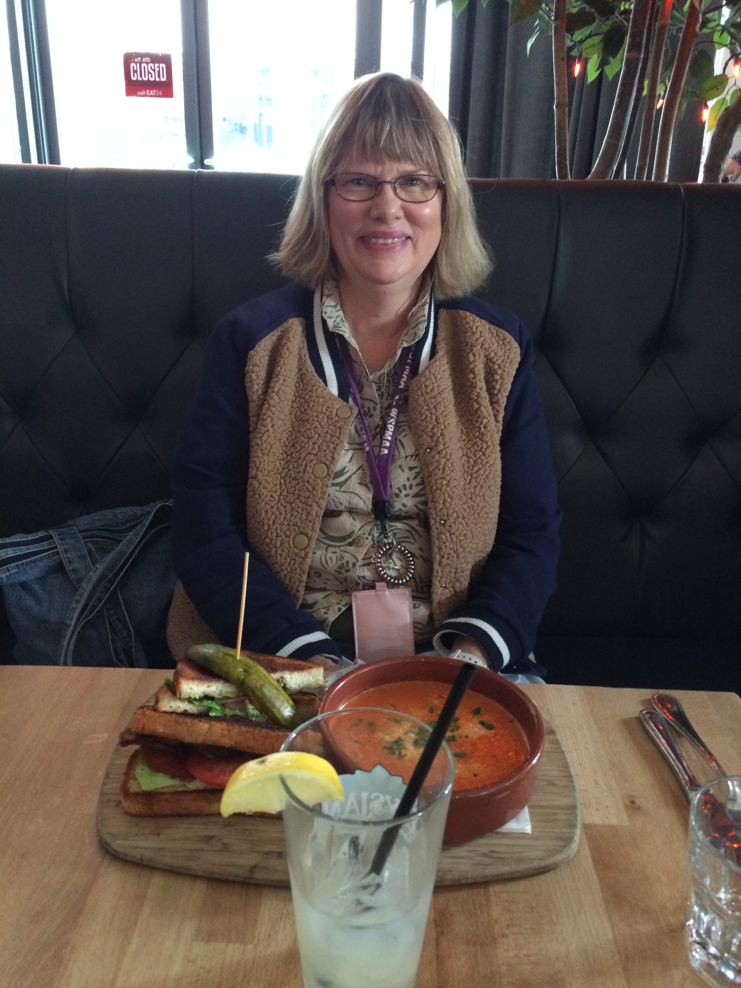 A woman with glasses and shoulder-length hair sits at a restaurant table with a grilled sandwich, a bowl of soup, and a glass of lemonade in front of her. She is smiling and wearing a jacket over a patterned shirt.
