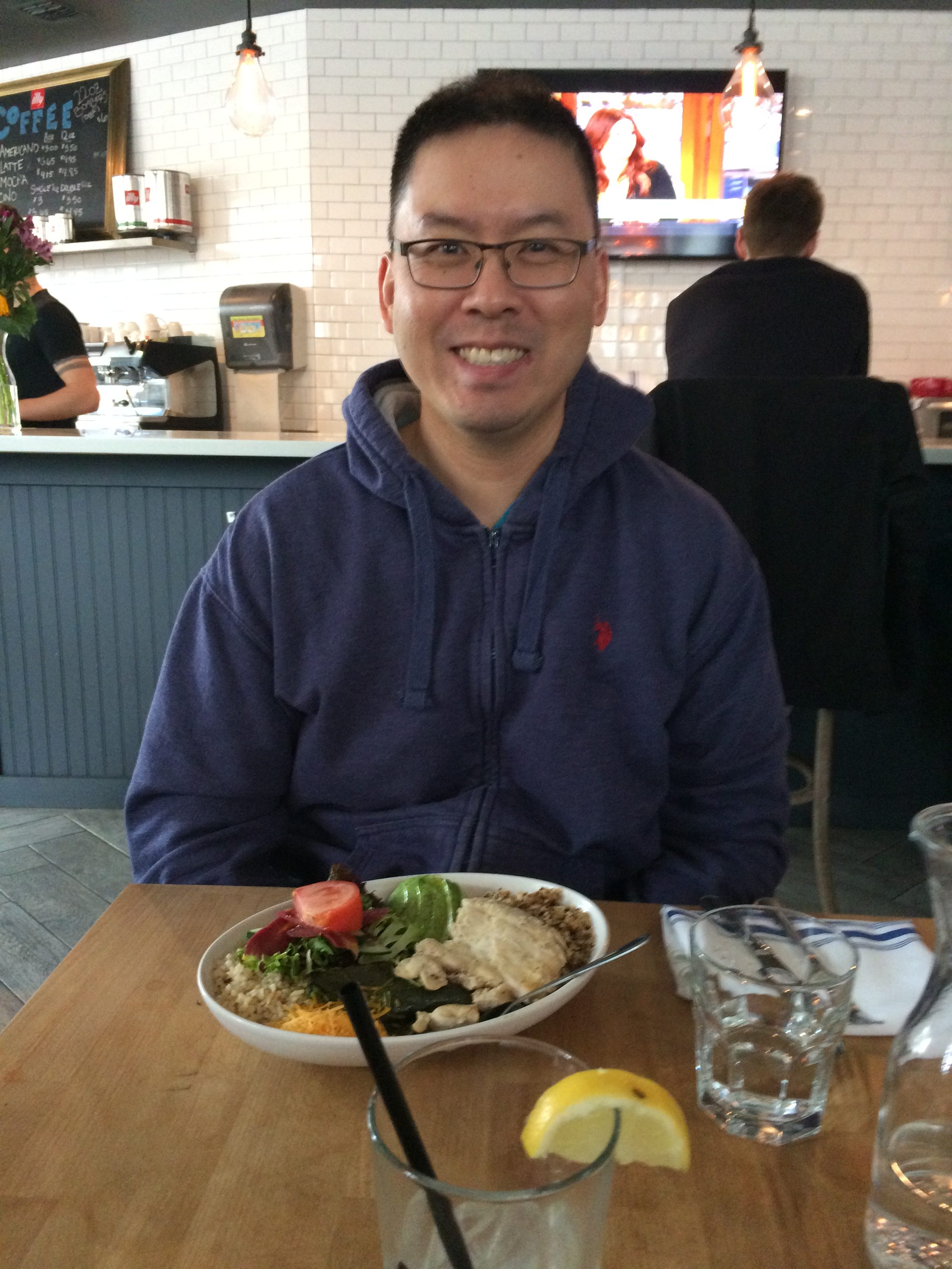 A man wearing glasses and a blue hoodie smiles while sitting at a table in a restaurant, with a plate of food in front of him. A TV and other diners are visible in the background.