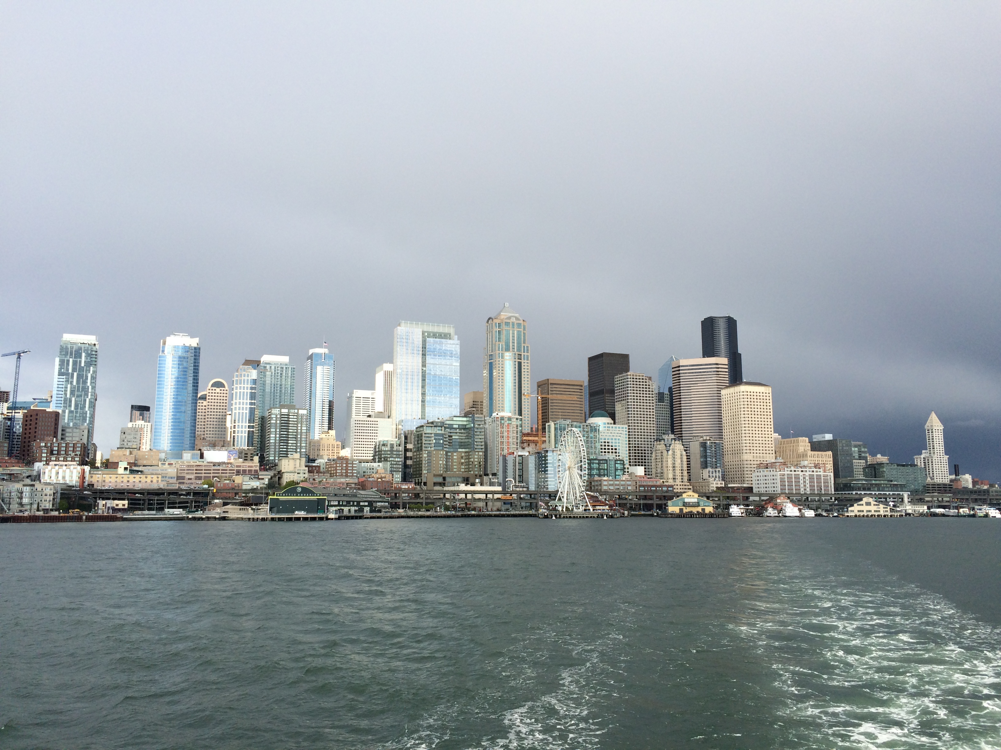 The image shows a daytime view of a city skyline with tall buildings along the waterfront under a cloudy sky. The water in the foreground reflects the buildings, and a Ferris wheel is visible near the shore.