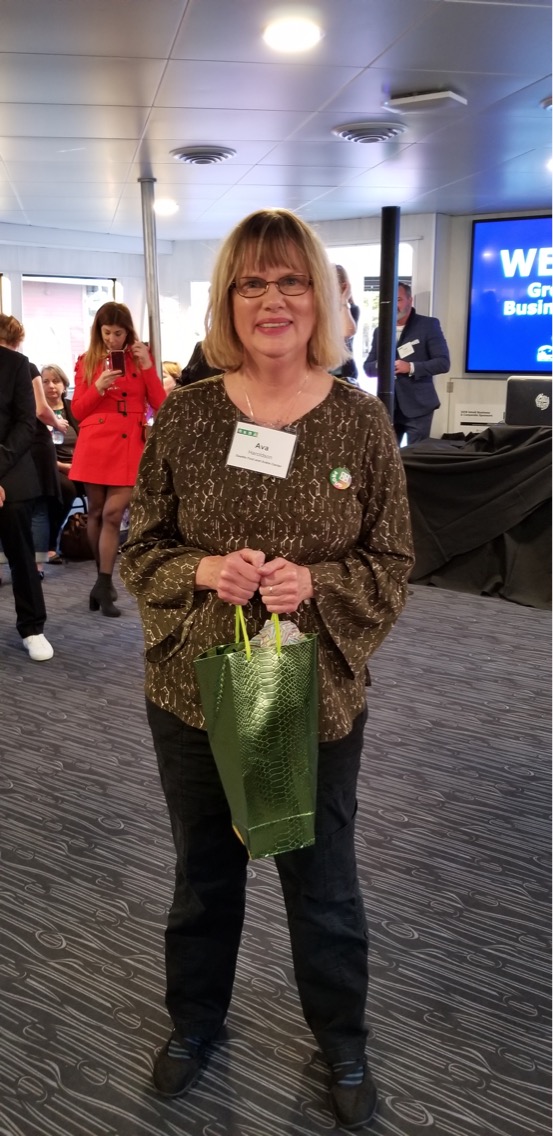 A woman with glasses and shoulder-length blonde hair stands indoors, smiling and holding a green gift bag. She wears a patterned top and nametag. People and a WE sign are visible in the background.