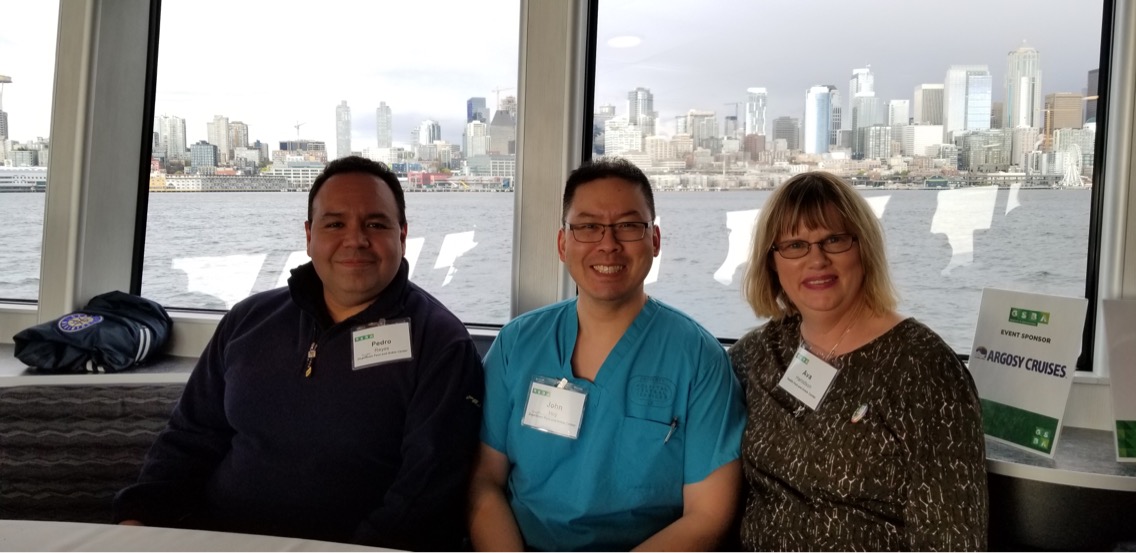 Three people sit and smile at a table on a boat with a city skyline and water visible through large windows behind them. They all wear name tags, and the atmosphere appears relaxed and friendly.