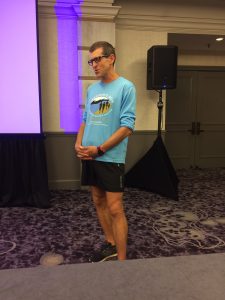 A man wearing glasses, a light blue long-sleeve shirt with a graphic, black shorts, and black sneakers stands indoors on a patterned carpet near a speaker and large screen.