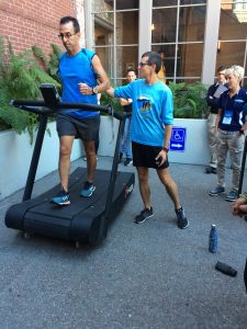 A man in athletic wear walks on a treadmill outdoors while another man in a blue shirt stands beside him, surrounded by onlookers. The scene takes place near a building with large windows and potted plants.
