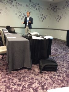 A man in a suit stands at the front of a conference room, gesturing while speaking. There are tables with gray tablecloths, a projector, papers, and a black bag on the purple-patterned carpet.