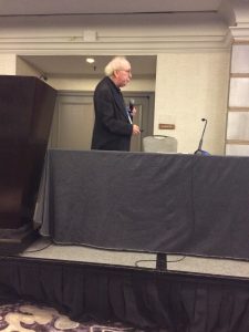 An older man with white hair and glasses stands holding a microphone next to a table and podium in a conference room, facing to the left.
