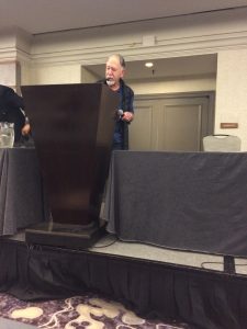 An older man stands behind a dark wooden podium on a stage, speaking into a microphone in a conference room. There is a table with a grey tablecloth and a pitcher of water beside him.