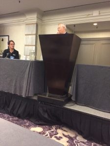 A man stands at a podium speaking into a microphone in a conference room, while a woman sits at a table nearby, listening. The tables are draped with gray cloths and a patterned carpet covers the floor.