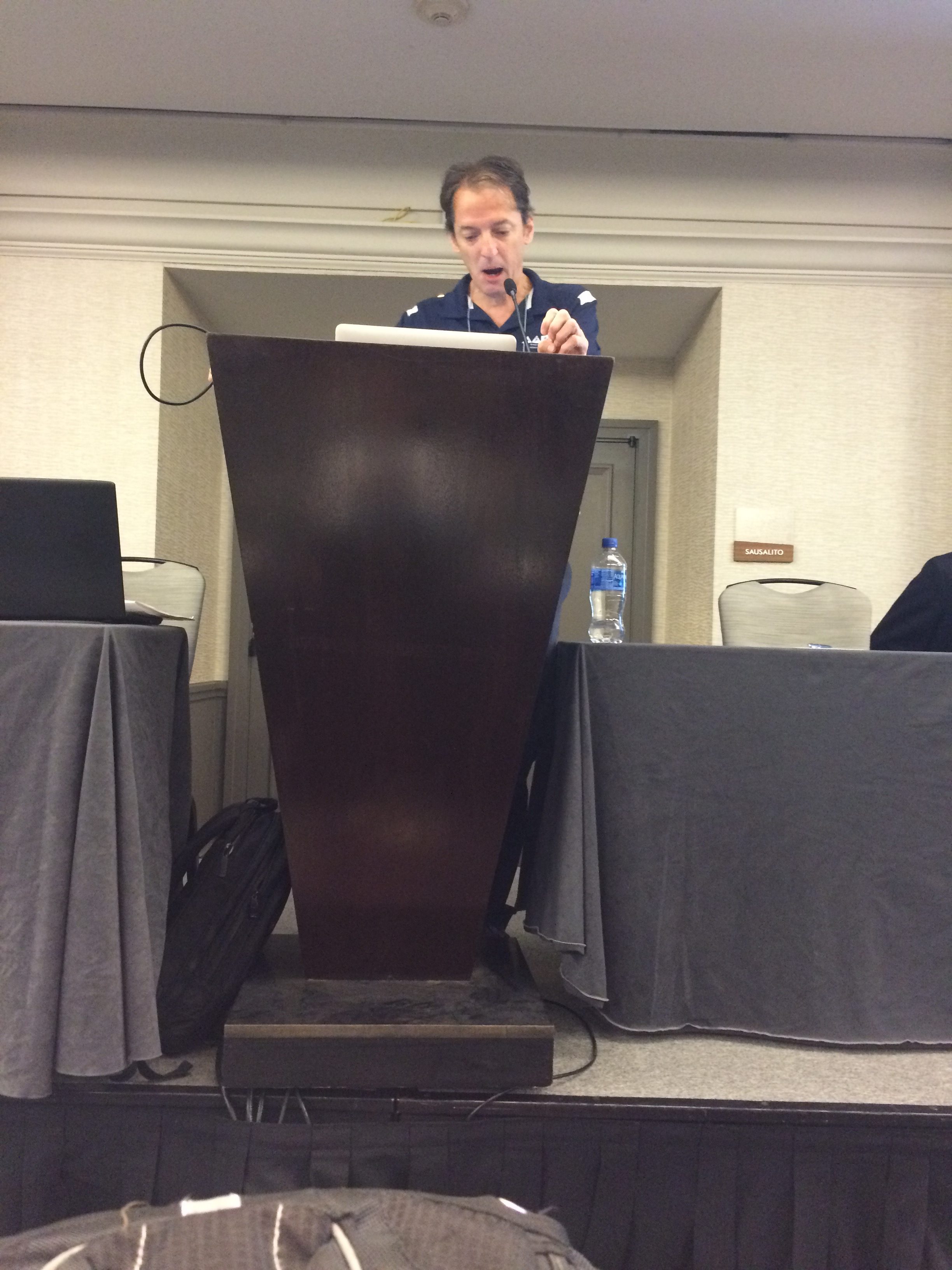 A man stands behind a tall wooden podium, speaking or presenting in a conference room. A laptop, water bottle, and chairs are visible on either side of him.