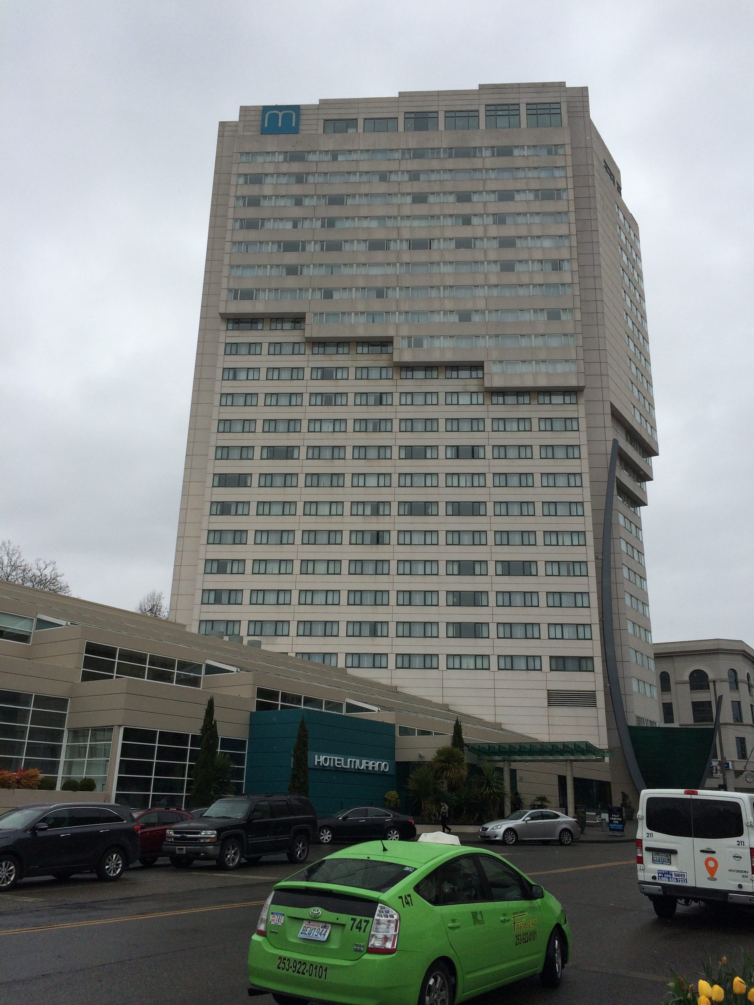 A tall, modern hotel building with many windows stands under a cloudy sky. A green taxi and several other vehicles are parked in front of the hotel entrance.