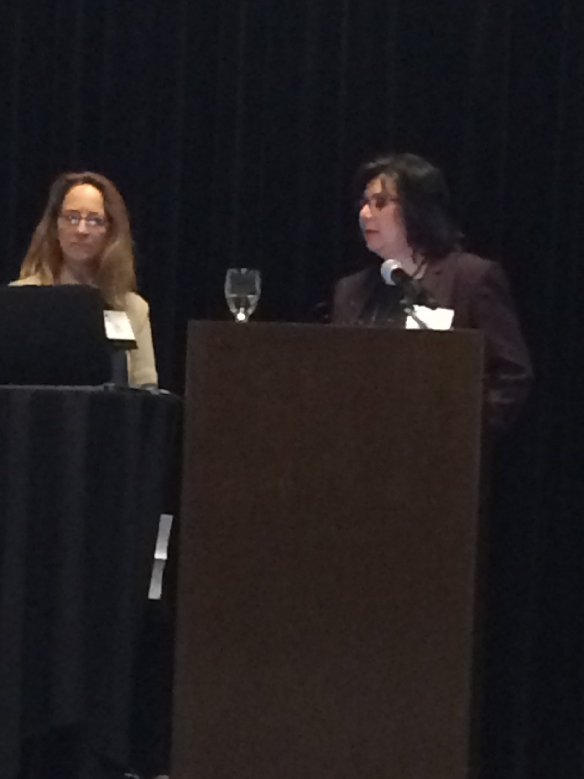 Two women stand behind podiums at a conference or formal event, one speaking into a microphone while the other listens. A glass of water sits on the podium. The background is dark and the lighting is dim.