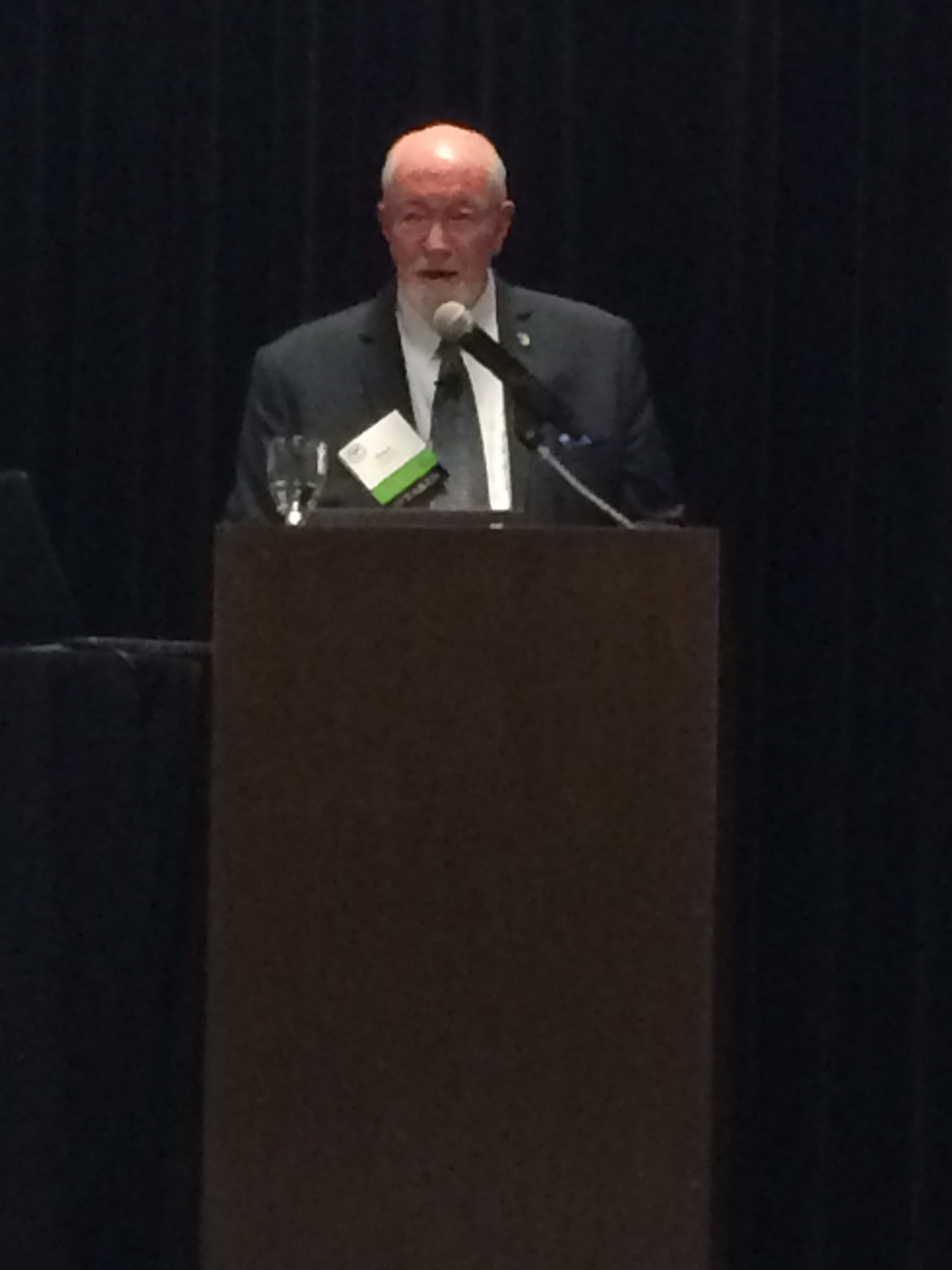 An older man with glasses and a beard stands at a podium, speaking into a microphone. He is dressed in a dark suit and tie, and wears a lanyard with name tags. The background is dark.