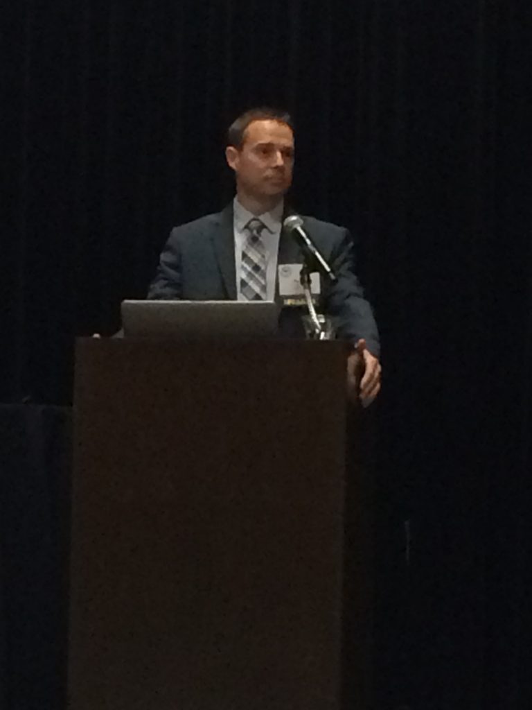 A man in a suit and tie stands behind a podium with a laptop, speaking at an event against a dark background.