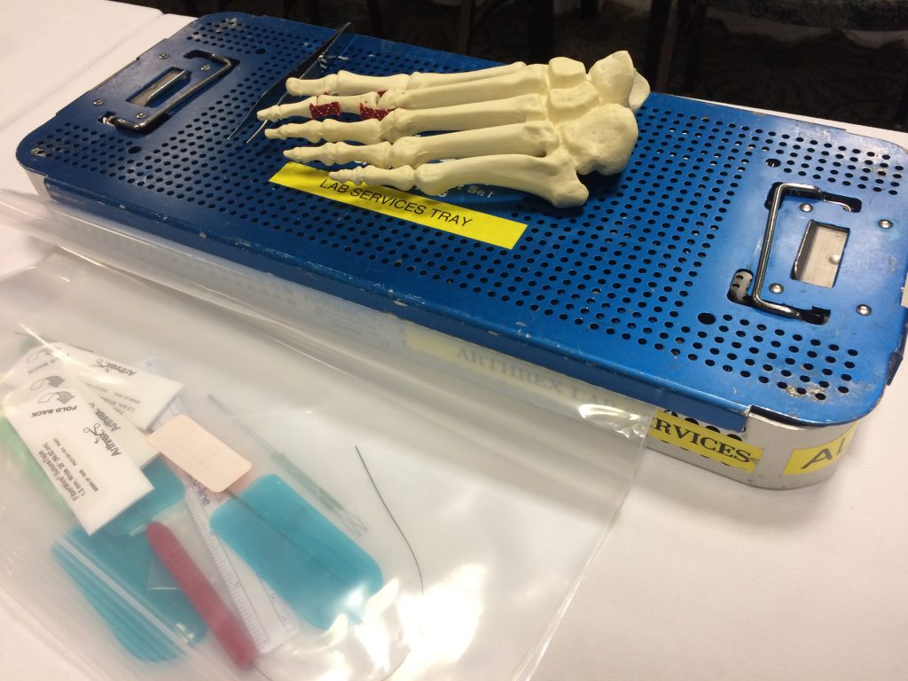 A blue metal surgical tray labeled “LAB SERVICES TRAY” holds a plastic model of a human foot. In front, a clear plastic bag contains medical tools, bandages, and surgical supplies.