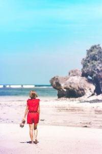 A woman in a red dress walks barefoot on a sandy beach, holding her shoes in one hand, with the ocean and large rocks visible in the background under a bright blue sky.