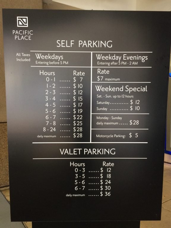 A sign displays parking rates at Pacific Place for self and valet parking, showing hourly and daily rates for weekdays, evenings, weekends, and motorcycles. It also lists special weekend and valet parking rates.