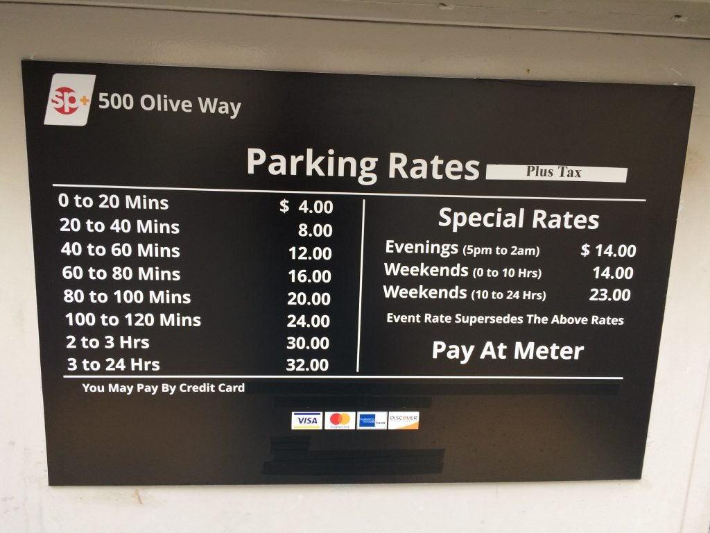 A sign displays parking rates for 500 Olive Way, listing prices by time intervals and special rates for evenings and weekends. It notes payment by credit card is accepted and special rates may override others.