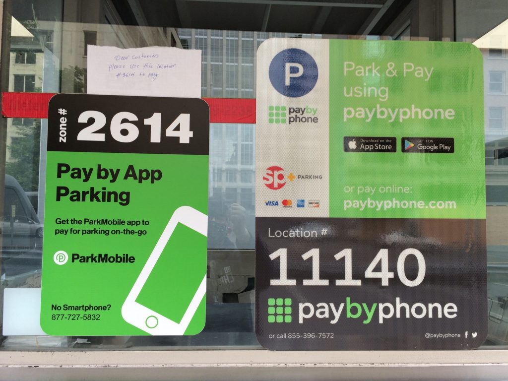 A window displays signs for mobile parking payment. One sign reads Zone #2614 Pay by App Parking, and another reads Park & Pay using paybyphone with location #11140 and payment instructions.