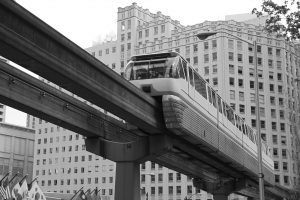 A monorail travels on an elevated track through a city with tall, rectangular buildings in the background. Flags are visible below, and the photo is in black and white.