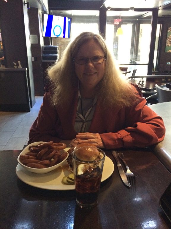 A woman with long blonde hair and glasses sits at a restaurant table with a plate of fries and a sandwich, and a glass of soda, smiling at the camera. The restaurant interior is dimly lit.