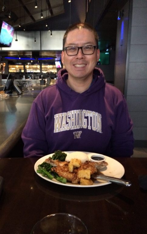A man wearing glasses and a purple Washington hoodie smiles while sitting at a restaurant table with a plate of food that includes greens and tofu in front of him.