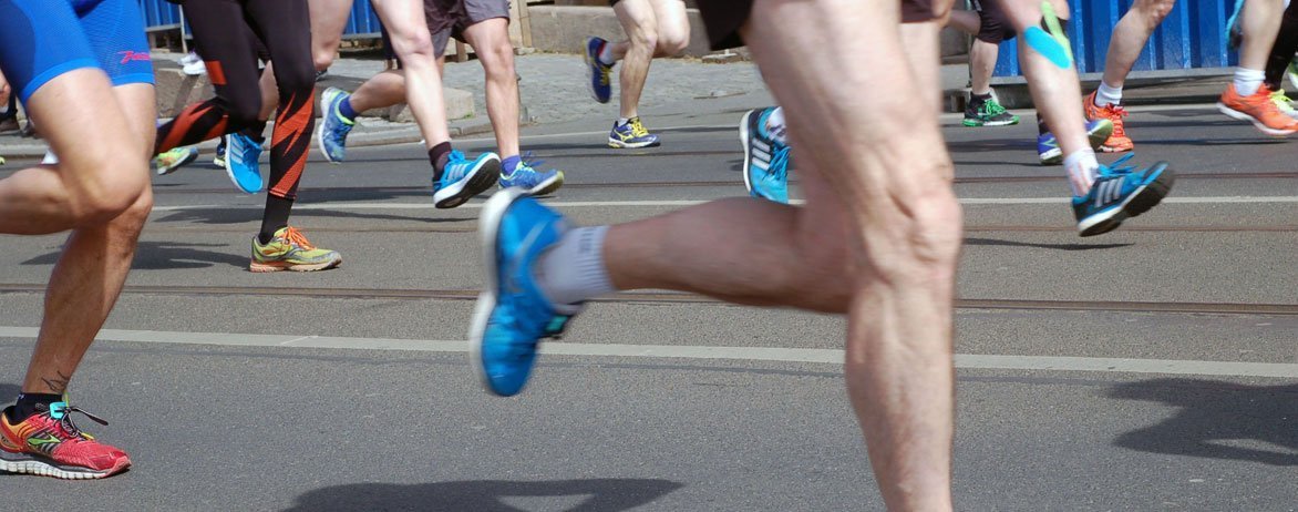 Runners in colorful athletic shoes and shorts run on a paved road during a race, with only their legs visible and the background slightly blurred.