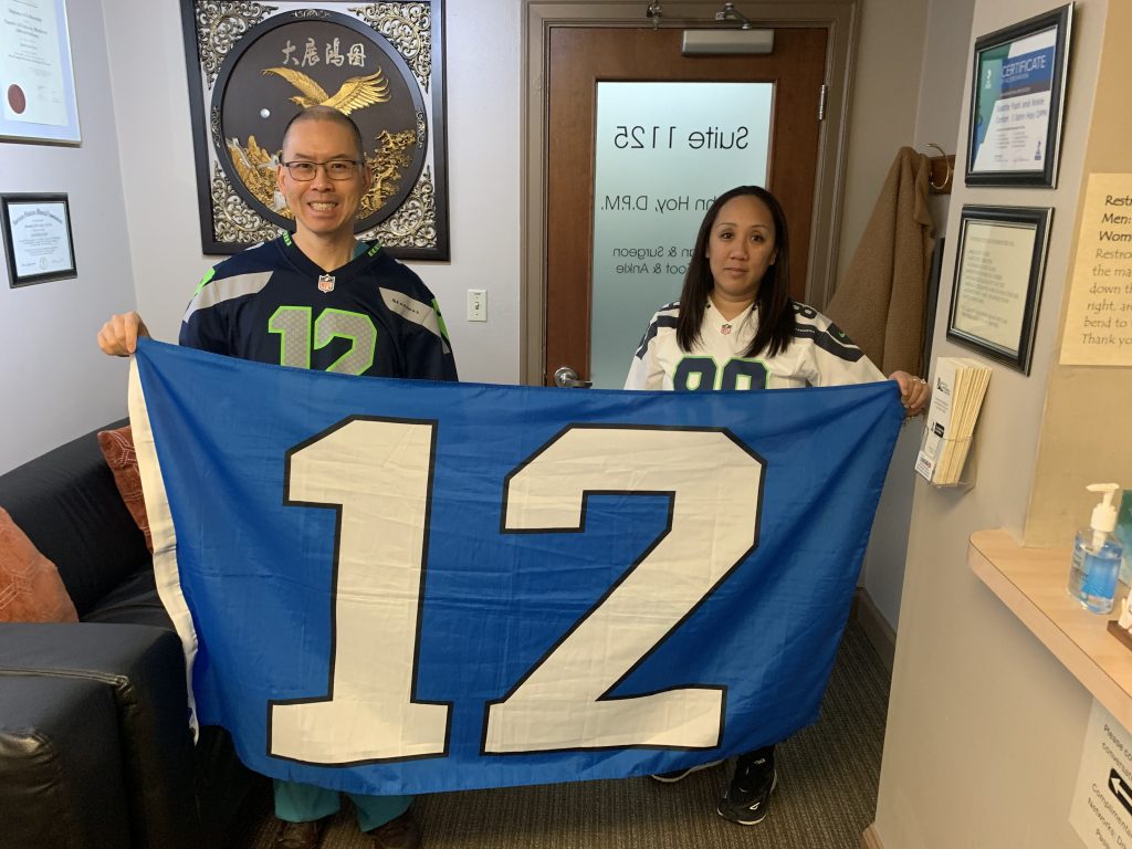 Two people wearing Seattle Seahawks jerseys hold a large blue, white, and gray “12” flag indoors. They are smiling, standing in an office with framed art and a door labeled “Suite 112.”.