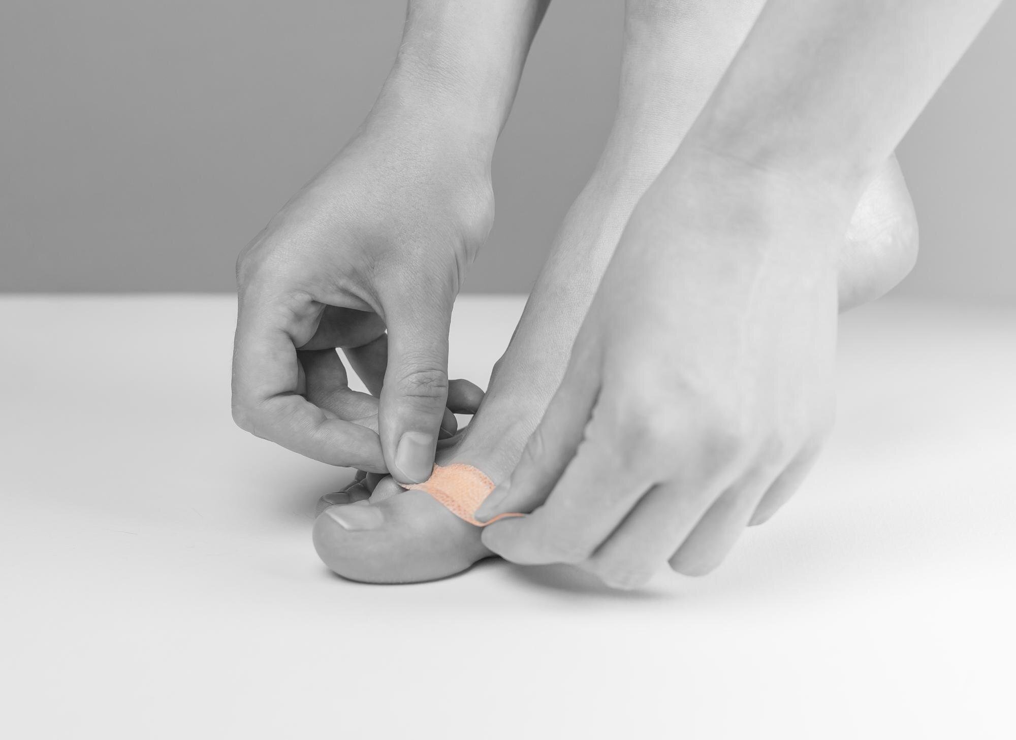 A person applies a small adhesive bandage to their big toe, with both hands gently pressing the bandage in place. The image is in black and white except for the colored bandage.