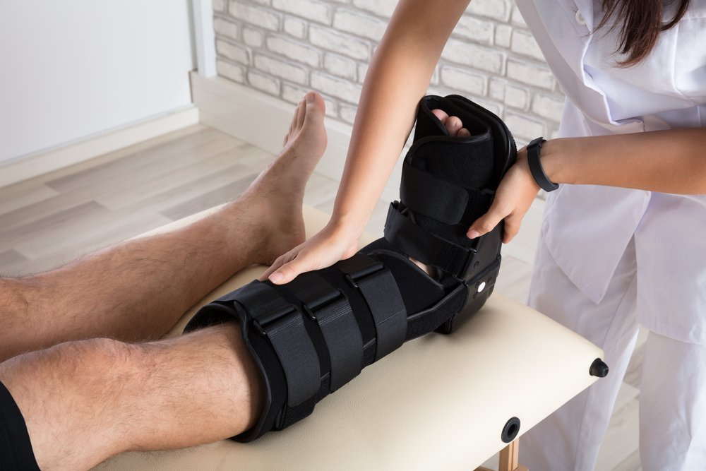 A medical professional helps fit a black orthopedic walking boot onto a persons lower leg and foot as the person lies on an exam table in a clinic.