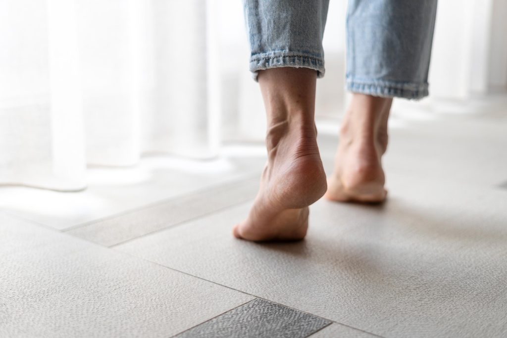 A person in rolled-up jeans walks barefoot on a light-colored floor near a window with sheer curtains, showing only their lower legs and feet.