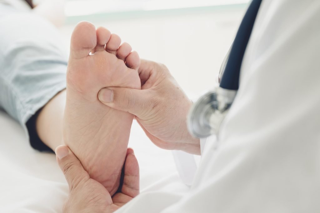 A doctor examines the bottom of a patients foot, gently pressing with both hands. The patient is lying down, and the doctors stethoscope is visible around their neck.