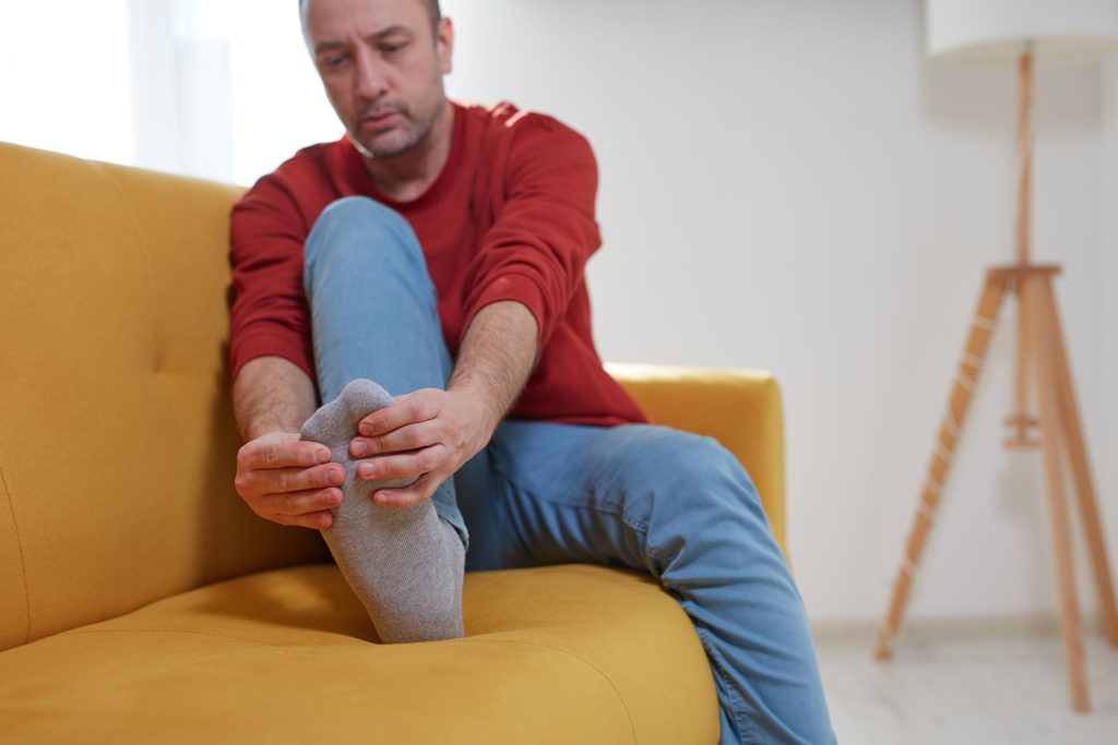A man in a red shirt and blue jeans sits on a yellow couch, holding his foot with both hands and appearing to be in pain or discomfort.