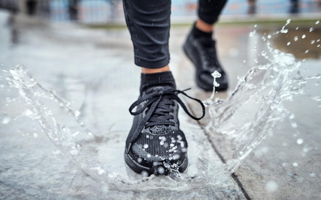 A person wearing black athletic shoes steps into a puddle, causing water to splash around their foot on a wet pavement.