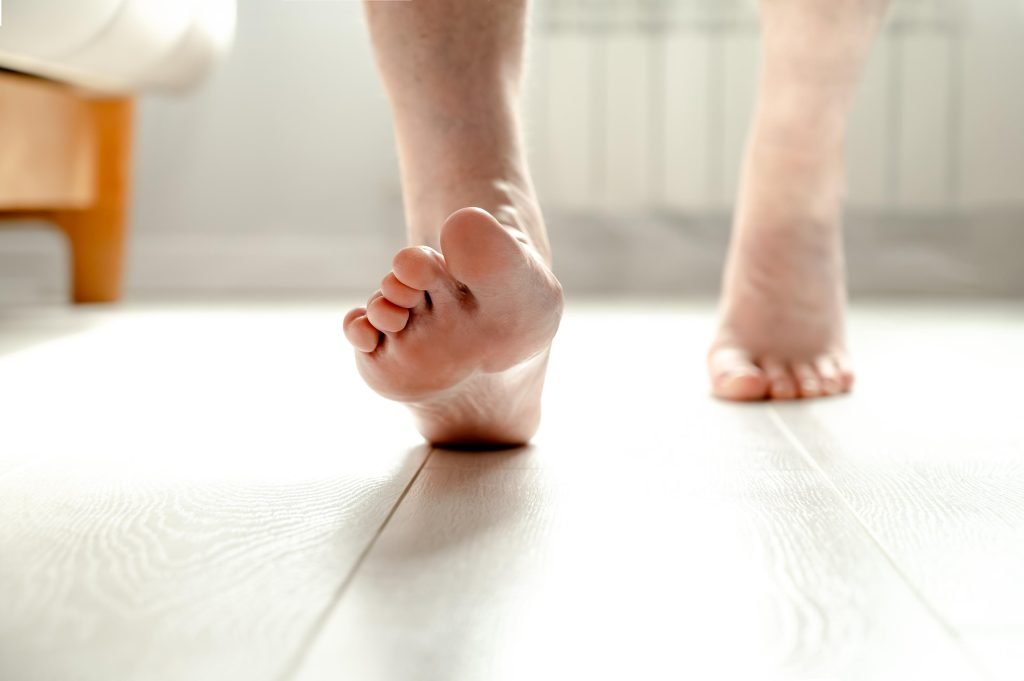 Close-up of bare feet walking on a light-colored wooden floor, with focus on the sole of the closest foot and soft natural light in the background.