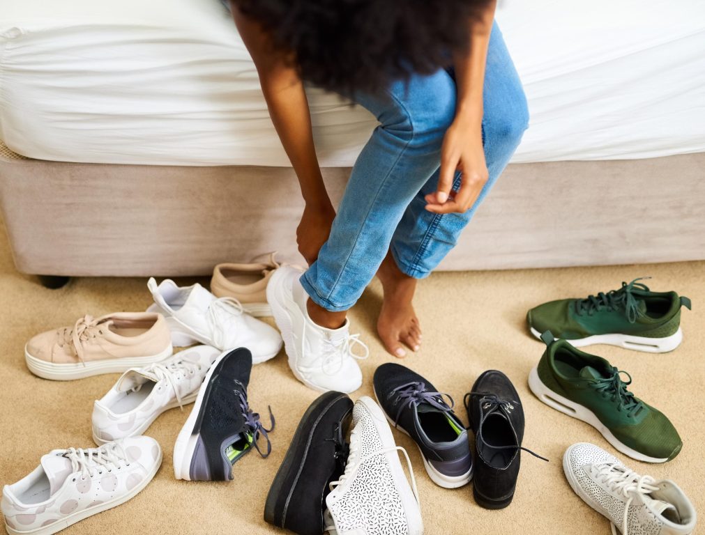 Person sitting on a bed, trying on white sneakers, surrounded by various pairs of shoes in different colors and styles on the floor.