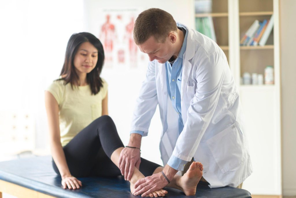 A doctor in a white coat examines a womans ankle as she sits on an exam table in a medical office. The woman appears relaxed and attentive during the checkup.