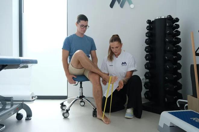 A woman in uniform helps a seated man stretch his foot using a yellow resistance band in a clinic room with gym equipment and weights.