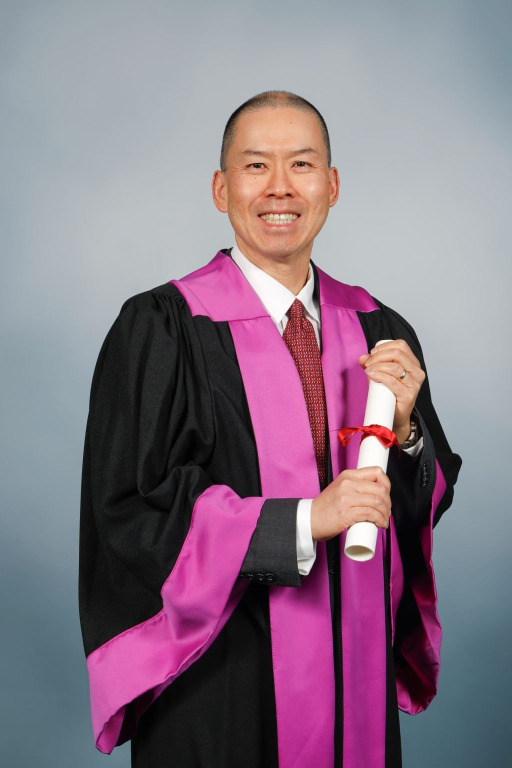 A person in a black graduation gown with purple trim smiles and holds a rolled-up diploma tied with a red ribbon, standing against a plain background.